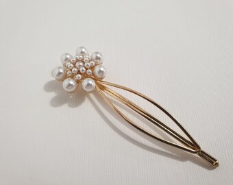 May include: A gold hair clip with a pearl flower design. The flower is made of small white pearls and is centered on the clip. The clip is long and thin, with a curved shape.