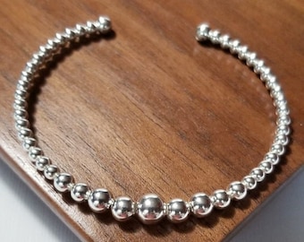 May include: A silver beaded bangle bracelet with a simple design. The bracelet is made of small, round beads that are evenly spaced.