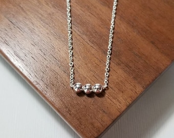 May include: A delicate silver necklace featuring three small, round beads. The necklace is displayed on a dark wooden surface, creating a contrast with the bright metal. The chain is fine and elegant.