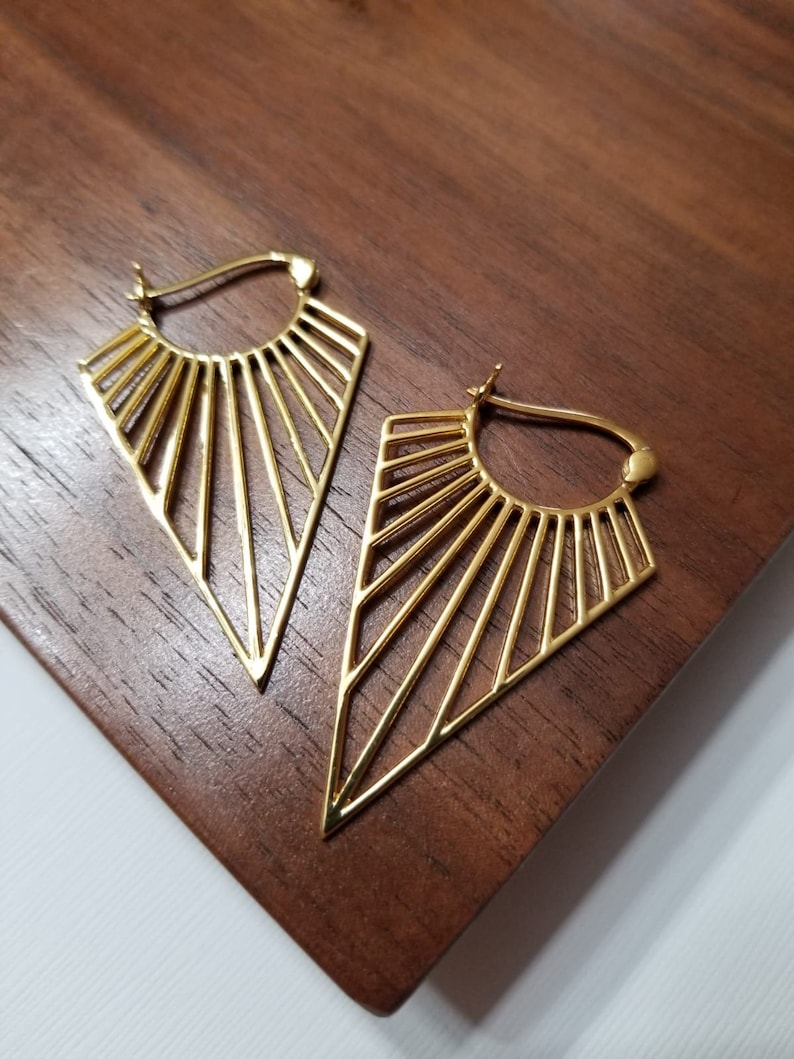 May include: A pair of gold-toned hoop earrings featuring a geometric, fan-like design with radiating thin metal bars that form a triangular shape. The earrings have a secure latch closure at the top and a polished, smooth metallic finish.