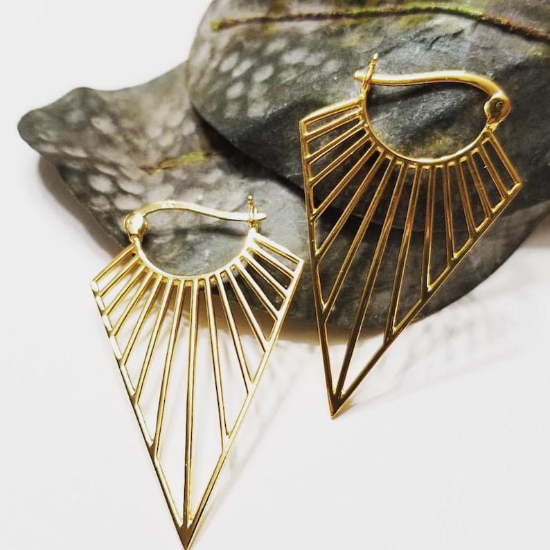 May include: A pair of gold-toned hoop earrings featuring a geometric fan-shaped design. Each earring consists of a curved top wire connected to a series of radiating thin metal bars that converge into a sharp, pointed triangular base.