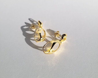 May include: A pair of gold-toned earrings with an abstract, organic design. The earrings are made of a smooth, shiny metal and have a dangling, teardrop shape.