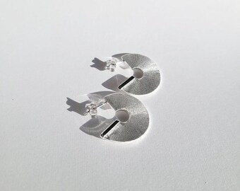 May include: A pair of silver hoop earrings with a textured finish. The earrings are in the shape of a half circle with a smaller circle cut out in the center.