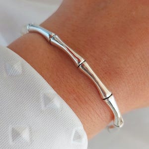 May include: A silver-colored bamboo-style bracelet. The bracelet is worn on a wrist, with a white textured shirt in the background. The bracelet has a segmented design, mimicking the look of bamboo stalks.
