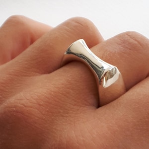 May include: A polished silver ring with a smooth, concave, hourglass-shaped top section that tapers toward the center. The band features a solid, reflective metallic surface with a curved design that sits across the finger.
