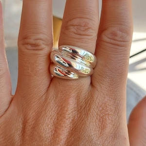 May include: A wide, polished silver ring featuring a multi-layered, twisted, or braided design. The band has a smooth, reflective metallic surface with rounded, overlapping segments that wrap around the finger, creating a bold, continuous, and sculptural appearance.