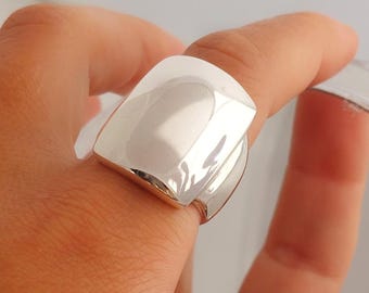 Puede incluir: Un anillo de plata pulida con una cara grande, lisa y cuadrada con esquinas redondeadas. La banda es ancha y refleja la luz en toda su superficie metálica, creando un aspecto brillante y reflectante en el dedo.