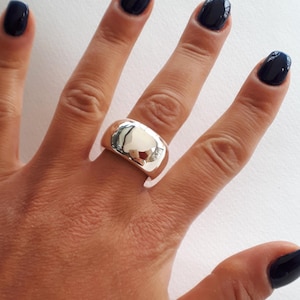 May include: A wide, polished silver-tone band ring with a smooth, reflective surface. The ring features a thick, rounded profile that wraps around the finger, showcasing a high-shine metallic finish that captures light and creates a mirror-like effect.