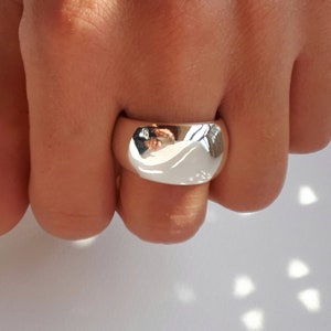 May include: A wide, polished silver-tone band ring with a smooth, rounded surface. The metal has a highly reflective finish that captures the surrounding light and environment, creating a bright, mirror-like appearance on the front of the ring.