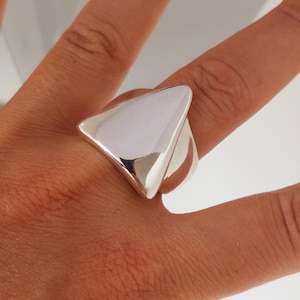 May include: A polished silver ring featuring a large, smooth, triangular-shaped face. The metal surface is highly reflective, showing a clean, solid finish that tapers into a wide band that wraps around the finger with a bold, geometric design.