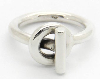 Può includere: Un anello d'argento con un designo a nodo unico. Il nodo è formato da un cerchio e una barra dritta che si incrociano.