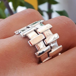 Peut inclure: Une bague en argent avec un design unique qui ressemble à un bracelet de montre. La bague est composée de plusieurs pièces rectangulaires reliées.