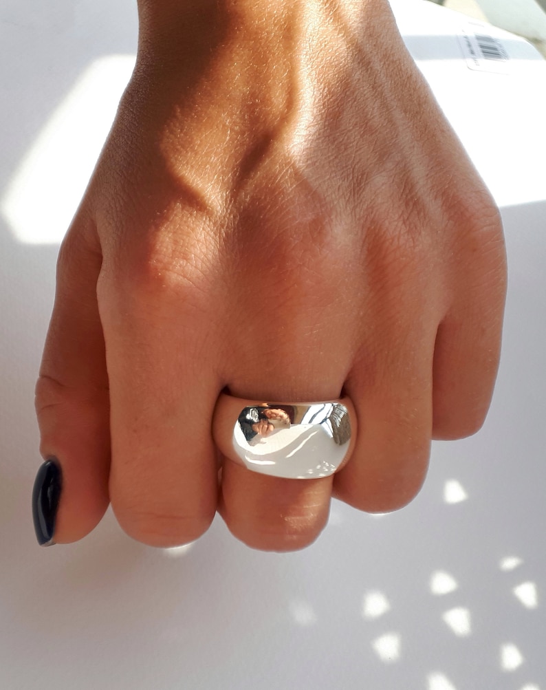 May include: A wide, polished silver-tone band ring with a smooth, rounded surface. The metal has a highly reflective finish that captures the surrounding light and environment, creating a bright, mirror-like appearance on the front of the ring.