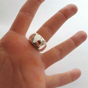 May include: A wide, polished silver-tone ring with a smooth, rounded, and reflective surface. The band features a thick, chunky design that sits prominently on the finger, showcasing a high-shine metallic finish that mirrors the surrounding environment.