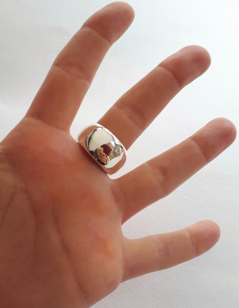 May include: A wide, polished silver-tone ring with a smooth, rounded, and reflective surface. The band features a thick, chunky design that sits prominently on the finger, showcasing a high-shine metallic finish that mirrors the surrounding environment.