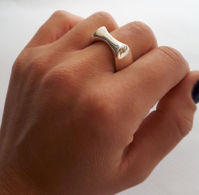 May include: A polished silver ring with a smooth, concave, hourglass-shaped top section that tapers toward the center. The band features a solid, reflective metallic surface with a curved design that sits across the finger.