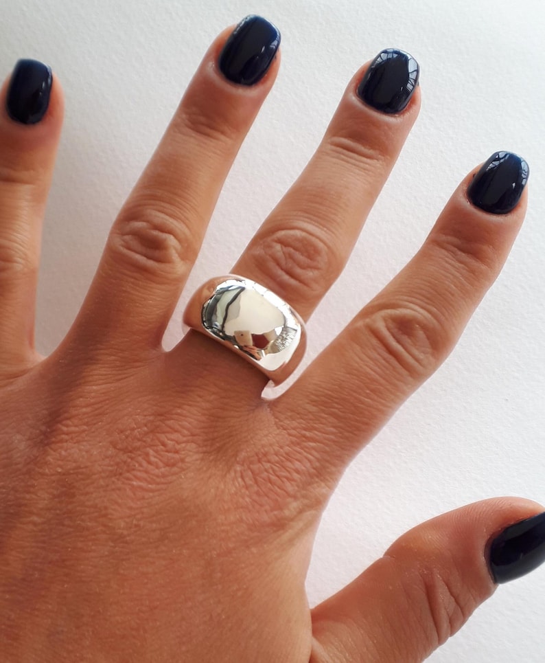 May include: A wide, polished silver-tone band ring with a smooth, reflective surface. The ring features a thick, rounded profile that wraps around the finger, showcasing a high-shine metallic finish that captures light and creates a mirror-like effect.