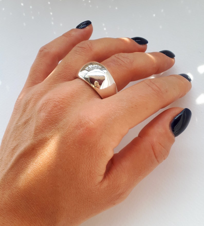 May include: A wide, polished silver-tone dome ring with a smooth, reflective surface. The band features a thick, rounded profile that tapers slightly toward the palm, creating a bold, solid appearance on the finger with high-shine metallic finish.