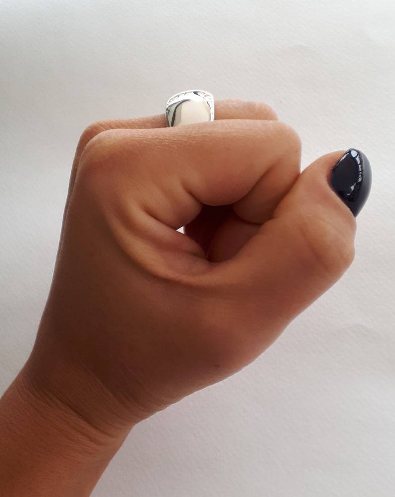 May include: A polished silver signet ring with a smooth, rounded rectangular face. The ring features a reflective metallic surface and is worn on the index finger, showcasing a clean, minimalist design with no engravings or additional embellishments.