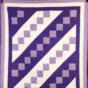May include: A purple and white quilt with a diagonal pattern of squares. The quilt is made with a variety of fabrics, including a floral print and a solid purple fabric.