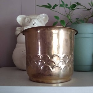 May include: A shiny, gold-colored metal planter with a scalloped pattern around the base. A white bear-shaped ceramic figure and a green potted plant are in the background. The planter is cylindrical and sits on a white shelf.