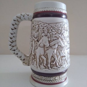 May include: White ceramic beer stein with a braided handle and a detailed relief scene of cowboys on horseback. The stein has a white lid and a base with the words "Cattle Drive" in a decorative border. The stein is approximately 6 inches tall.