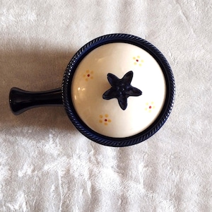 May include: A ceramic serving dish with a lid and handle. The dish is white with a navy blue star and floral accents. The rim and handle are navy blue. The dish is approximately 6 inches in diameter.