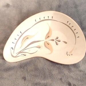 May include: Cream-colored, kidney-shaped dish with a floral design. The dish features a central flower motif in tan and gray, with matching accents along the rim. The design is reminiscent of mid-century modern pottery.