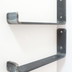 May include: Two black metal shelf brackets with a curved front edge. The brackets are mounted to a white wall.