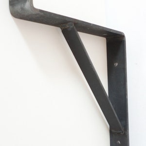 May include: A black metal shelf bracket with a triangular support. The bracket is mounted to a white wall.