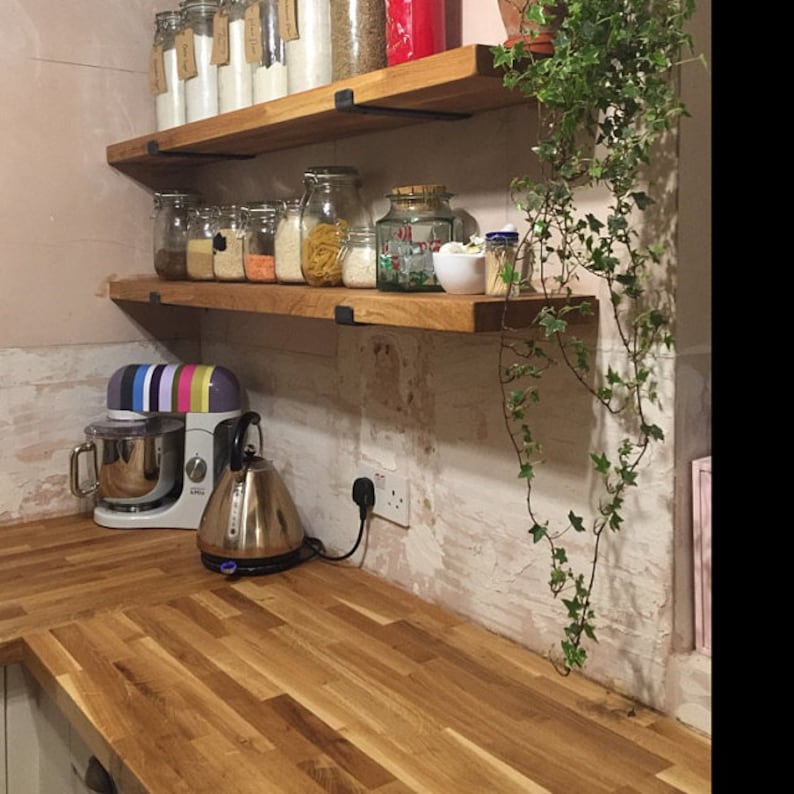 May include: Two wooden shelves with black metal brackets, mounted on a white tiled wall. The shelves are filled with glass jars containing various dry goods, such as pasta, rice, and flour. A potted plant with green leaves hangs from the top shelf.