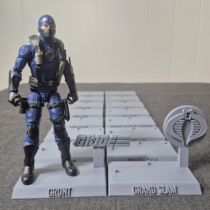 May include: A G.I. Joe action figure in a blue and black uniform stands on a gray platform. The platform has the word "GRUNT" printed on it. The G.I. Joe logo and the words "GRAND SLAM" are also visible on other platforms.