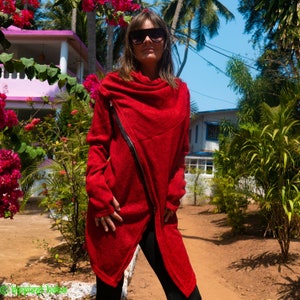 May include: A woman wearing a red, asymmetrical zippered jacket with a cowl neck. She is standing in front of a house with lush greenery.