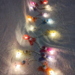 May include: A string of fairy lights with butterfly-shaped bulbs. The bulbs are in various colors, including pink, blue, yellow, and green. The lights are strung on a clear wire and are lit up, casting a warm glow.