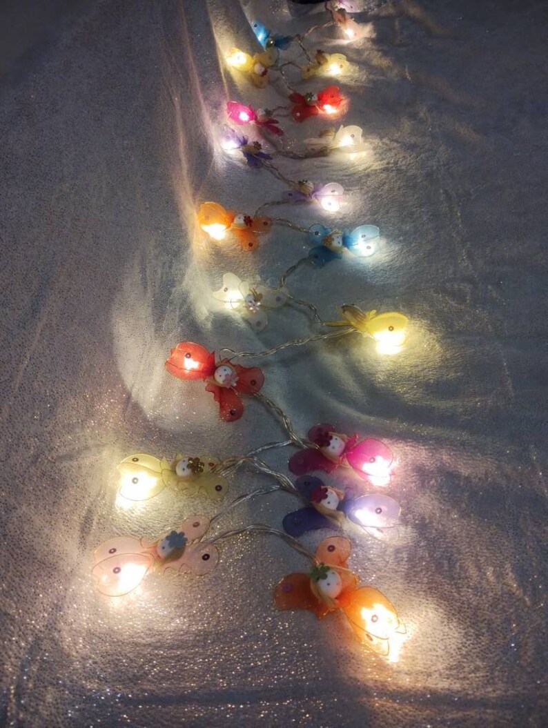 May include: A string of fairy lights with butterfly-shaped bulbs. The bulbs are in various colors, including pink, blue, yellow, and green. The lights are strung on a clear wire and are lit up, casting a warm glow.