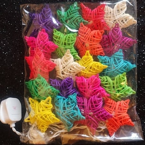 Könnte beinhalten: Eine Lichterkette mit geflochtenen sternförmigen Glühbirnen in verschiedenen Farben, darunter Weiß, Rosa, Blau, Grün, Gelb, Orange und Lila. Die Lichter sind in einem durchsichtigen Plastikbeutel mit einem weißen Stecker verpackt.