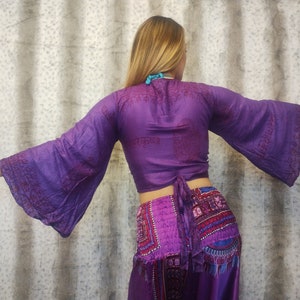 Könnte beinhalten: Lilafarbenes Seidenoberteil mit langen Glockenärmeln und einem Bindeband in der Taille. Das Oberteil hat ein bedrucktes Design mit Sanskrit-Text.