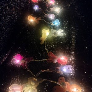May include: A string of fairy lights shaped like butterflies in various colors, including pink, purple, blue, yellow, and orange. The lights are strung on a clear wire and are lit up, creating a warm and festive atmosphere.