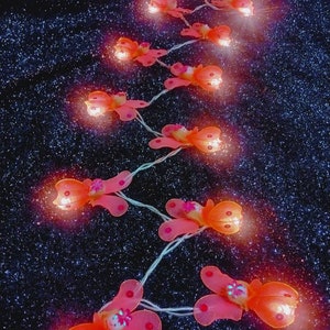 Puede incluir: Una cadena de luces con bombillas en forma de mariposa. Las luces son de color naranja y tienen un brillo cálido. Las luces están enhebradas en un cable transparente y están colgadas sobre un fondo oscuro.