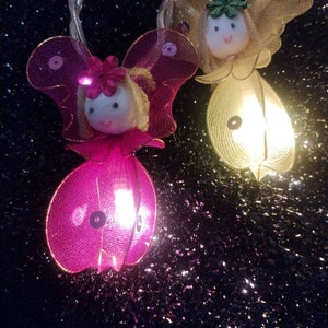 May include: A string of fairy lights with pink and yellow fairy figures. Each fairy has a light inside and is made of mesh fabric. The fairies have wings and faces with painted features.