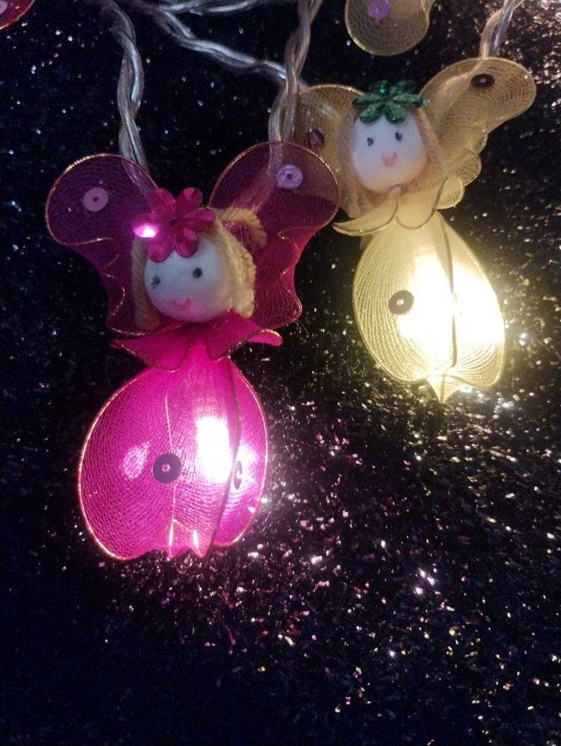 May include: A string of fairy lights with pink and yellow fairy figures. Each fairy has a light inside and is made of mesh fabric. The fairies have wings and faces with painted features.