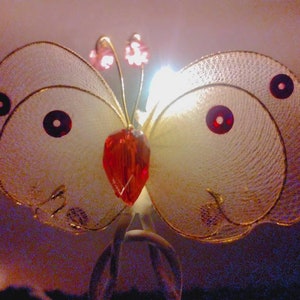 May include: A delicate butterfly-shaped ornament with a red crystal center and gold wire wings. The wings are translucent and have a subtle shimmer.