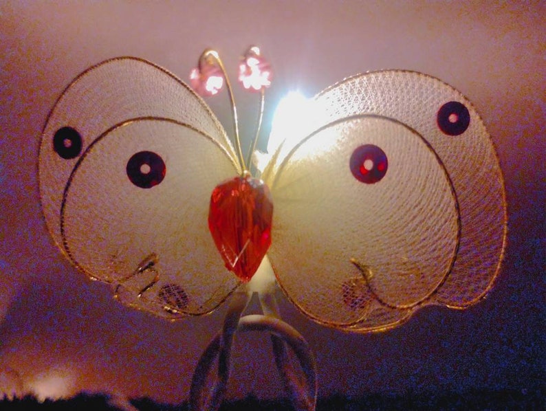May include: A delicate butterfly-shaped ornament with a red crystal center and gold wire wings. The wings are translucent and have a subtle shimmer.