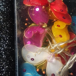 May include: A string of fairy lights with butterfly-shaped bulbs. The butterflies are made of sheer fabric and come in a variety of colors, including red, pink, yellow, blue, and white. Each butterfly has a small, clear crystal in the center.