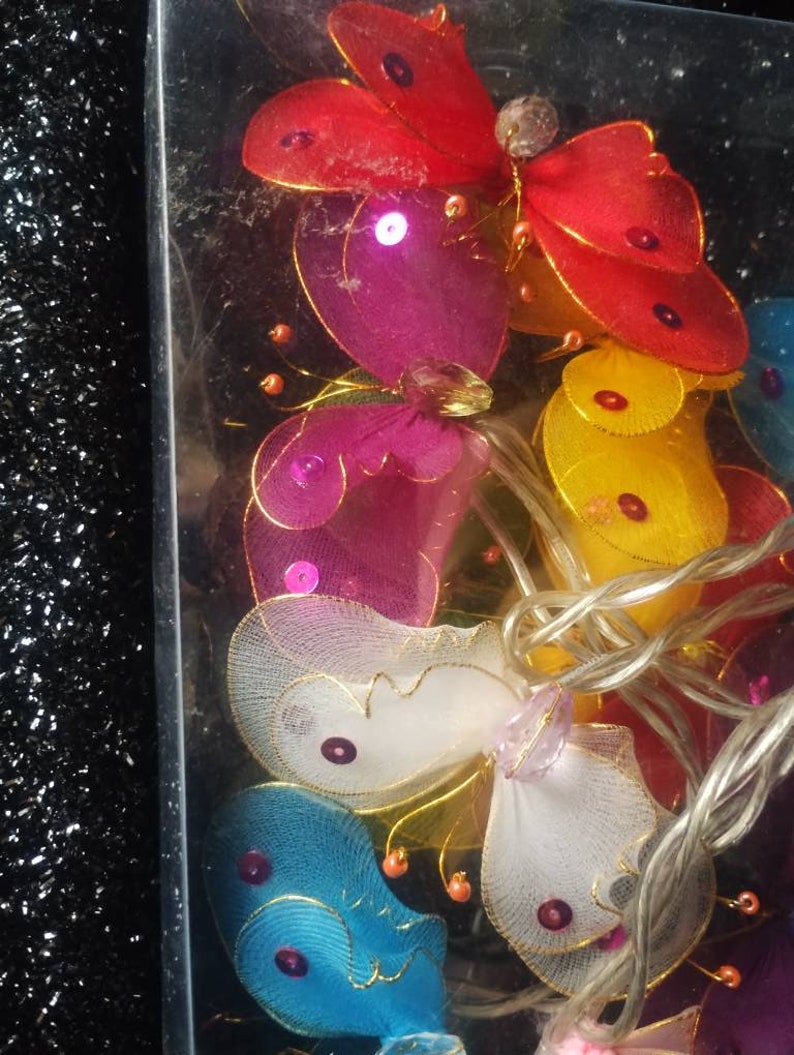 May include: A string of fairy lights with butterfly-shaped bulbs. The butterflies are made of sheer fabric and come in a variety of colors, including red, pink, yellow, blue, and white. Each butterfly has a small, clear crystal in the center.