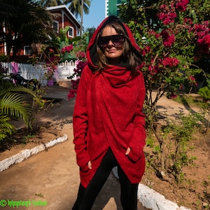 Può includere: Una donna indossa un maglione rosso con cappuccio e fodera nera. Il maglione ha uno stile avvolgente e un design lungo e fluido. Indossa pantaloni neri e occhiali da sole.