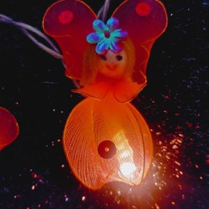 May include: A small orange and red fairy light with a doll-like face and a blue flower on its head. The light is on and glowing.
