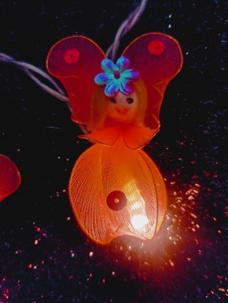 May include: A small orange and red fairy light with a doll-like face and a blue flower on its head. The light is on and glowing.