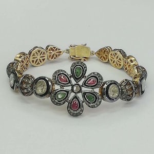 Könnte beinhalten: Ein Armband mit einem zentralen Blumenmotiv aus abwechselnd tropfenförmigen grünen und rosa Steinen, umgeben von kleinen Pavé-Steinen. Das Band besteht aus kreisförmigen Gliedern mit filigranen Metallarbeiten und einem goldenen Kastenschloss.