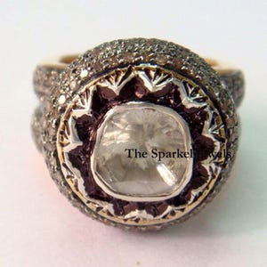 Può includere: Un anello d'argento con una grande pietra preziosa quadrata trasparente circondata da diamanti più piccoli. L'anello ha un design decorativo e ornato ed è incastonato su una fascia d'oro. Il testo "The Sparkle Jewels" è visibile sull'anello.